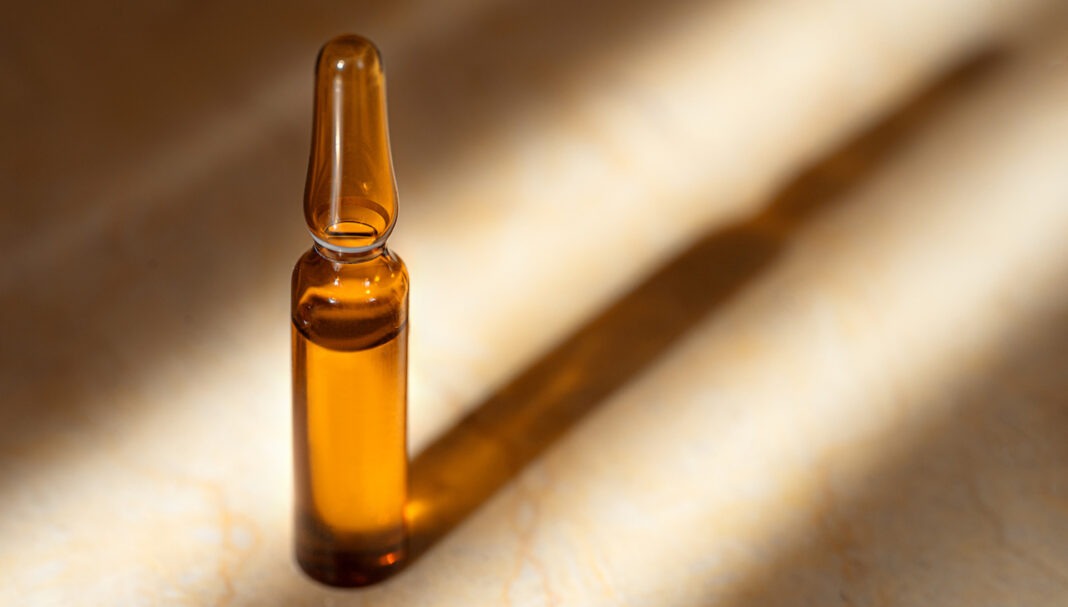 Glass ampoule of the Cosmetic product with natural origin Hyaluronic Acid and Vitamin C on a beige marble background with shadow.