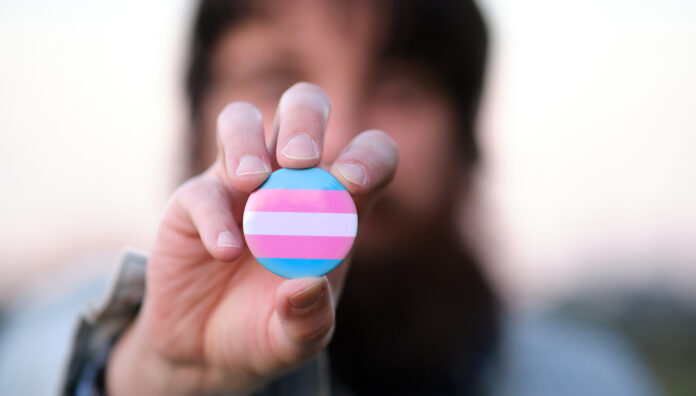 An unrecognizable bearded man holding and showing a pin with transgender colors flag