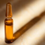 Glass ampoule of the Cosmetic product with natural origin Hyaluronic Acid and Vitamin C on a beige marble background with shadow.